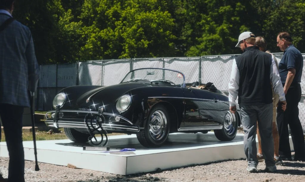 A black Porsche 356 is displayed on a pedestal with spectators nearby at a car event.