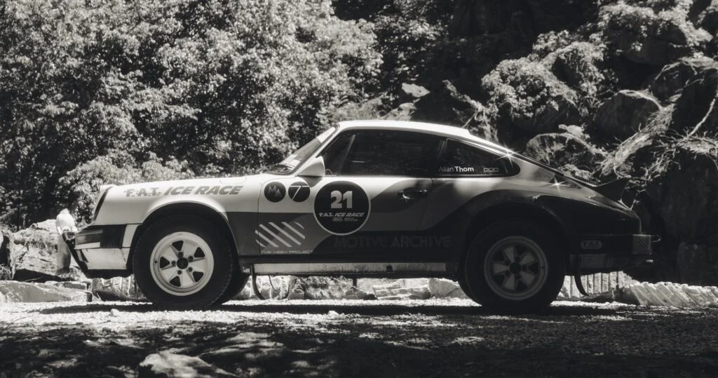 A black and white image of a Porsche 911 rally car parked on gravel in a forested setting.