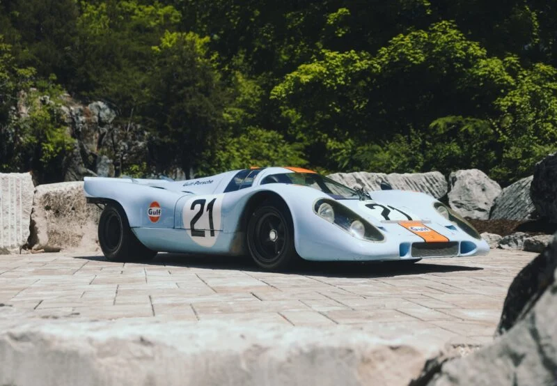 Porsche 917 race car in a light blue color with orange accents parked on a stone surface surrounded by greenery.