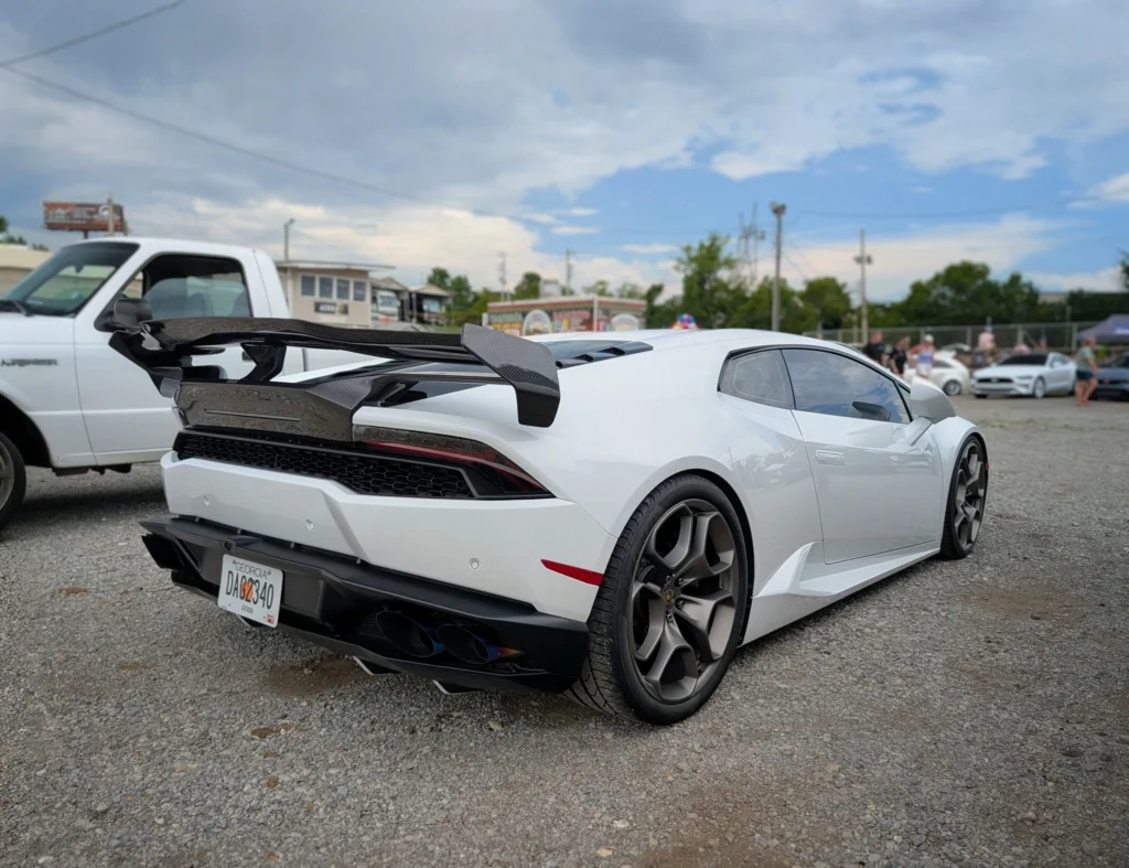 White Lamborghini Huracan with black carbon fiber rear wing at a car event on gravel with other vehicles and people in the ba