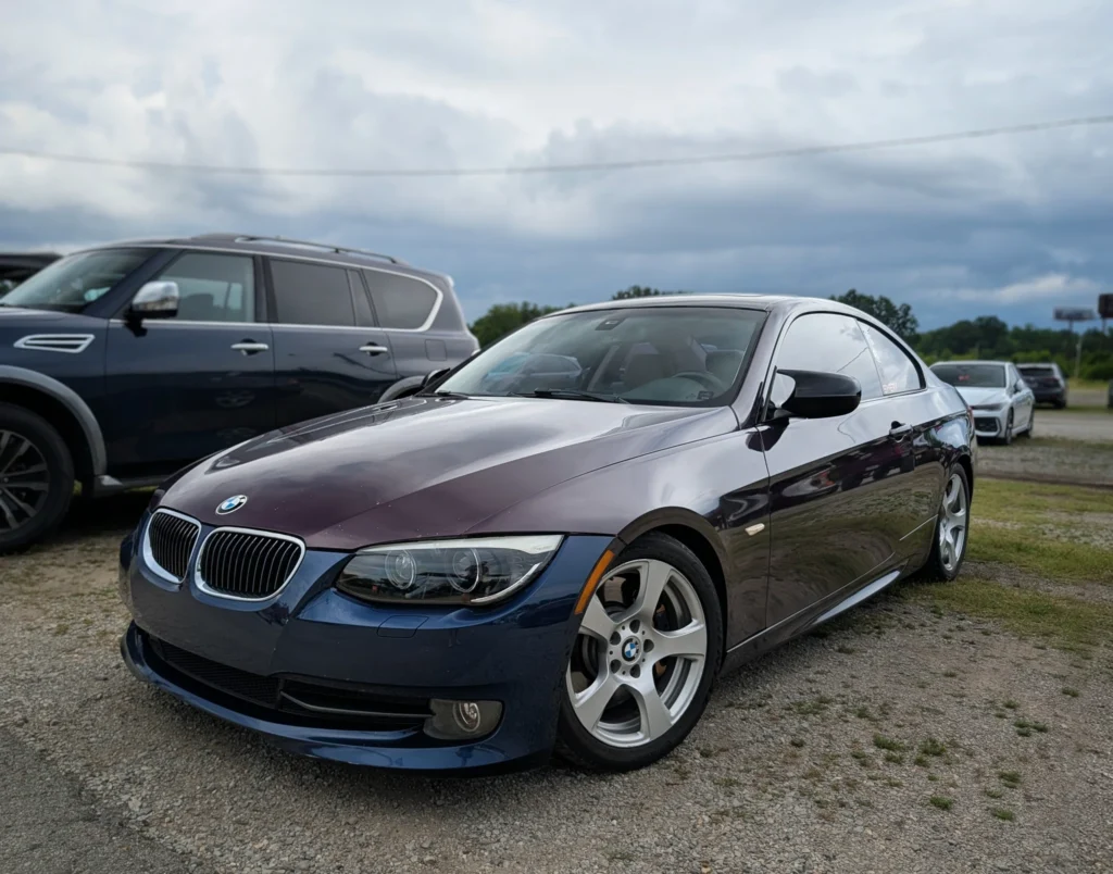 Dark blue BMW 3 series parked on gravel with other vehicles nearby under a cloudy sky