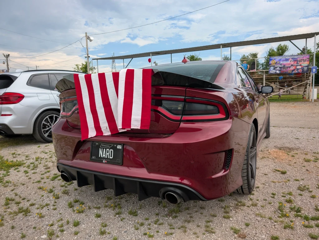 Rear view of a maroon Dodge Charger with an American flag draped over the spoiler at an outdoor event with bleachers and a wh