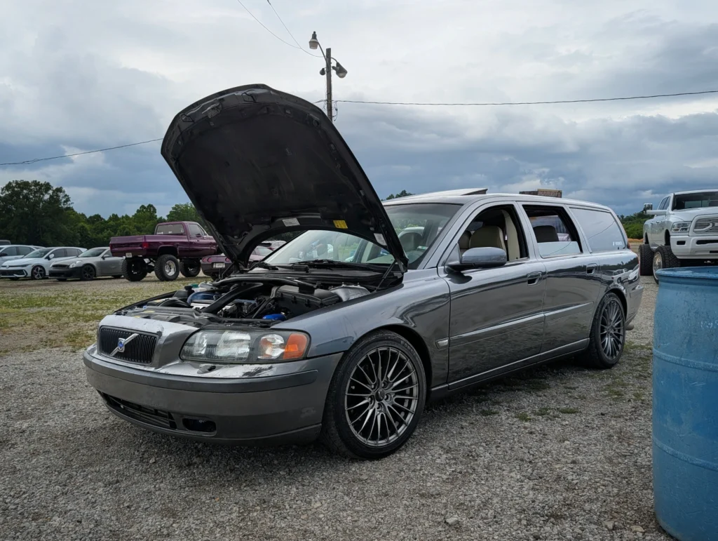 Gray Volvo V70 wagon with open hood parked on gravel with other vehicles and trees in the background under cloudy sky