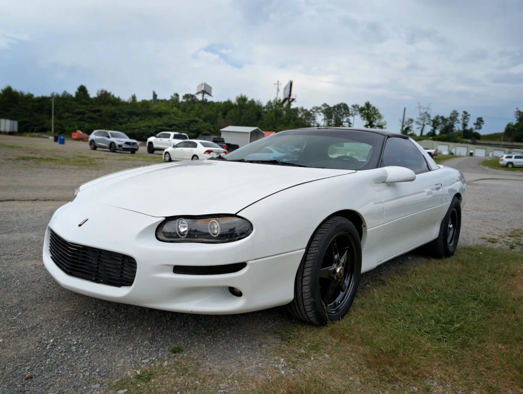 White Chevrolet Camaro with black wheels parked on gravel and grass at a car event with other vehicles and trees in backgroun