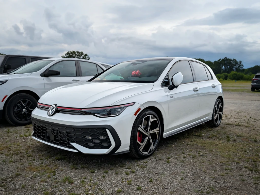 White Volkswagen GTI parked on gravel with other vehicles and a cloudy sky in the background