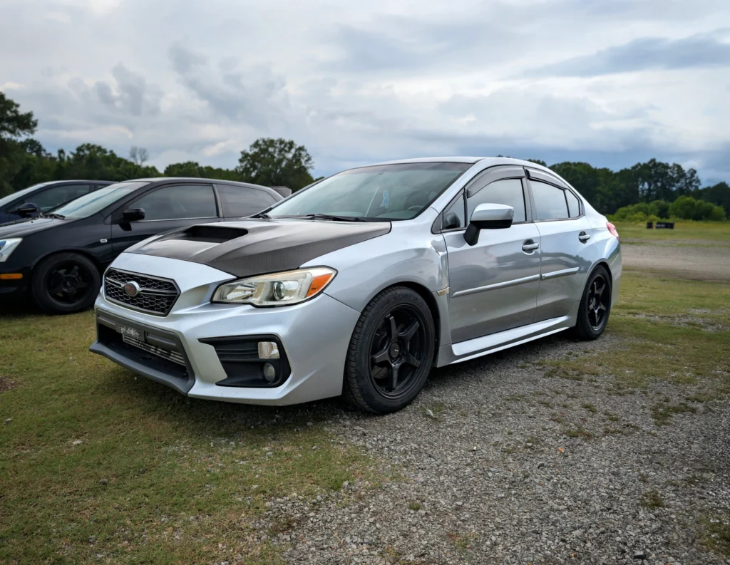 Silver Subaru WRX with black aftermarket hood and black wheels parked on grass with other cars and trees in background