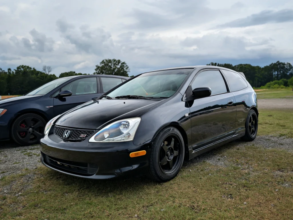 Black Honda Civic Si hatchback parked on grass with a gray Volkswagen GTI beside it under a cloudy sky