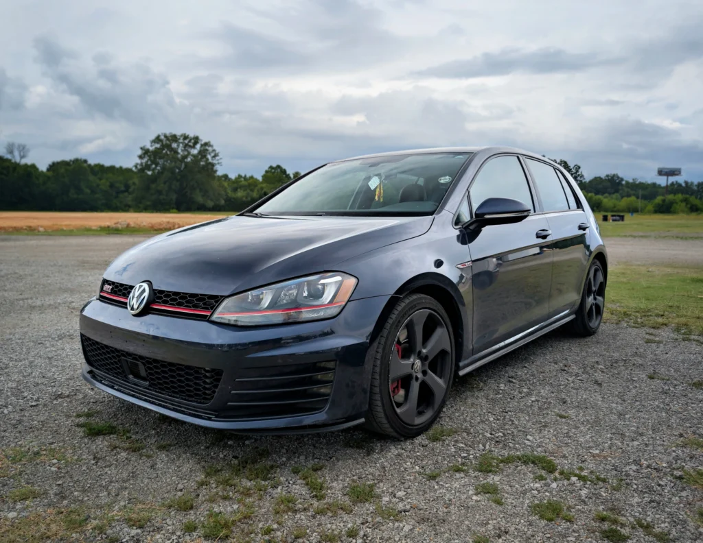 Black Volkswagen Golf GTI parked on a gravel area with trees and cloudy sky in the background