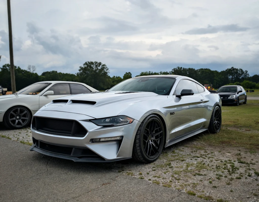 Silver Ford Mustang 5.0 parked on gravel and grass with other cars behind under a cloudy sky