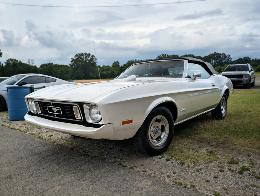 White Ford Mustang convertible parked on grass and pavement at an outdoor car event with other cars and trees in the backgrou