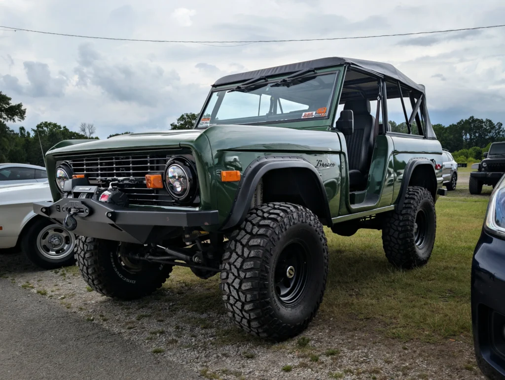 Green Ford Bronco with black wheels and an open soft top parked on a gravel lot with other vehicles nearby