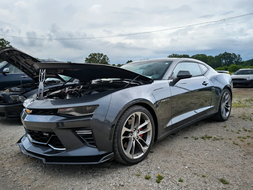 Black Chevrolet Camaro SS with hood open at an outdoor car event with other vehicles and trees in the background