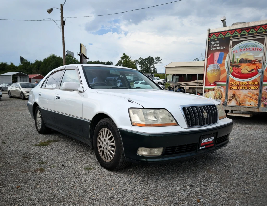 Toyota Crown Majesta two-tone parked on gravel near food truck with taco signage under cloudy sky