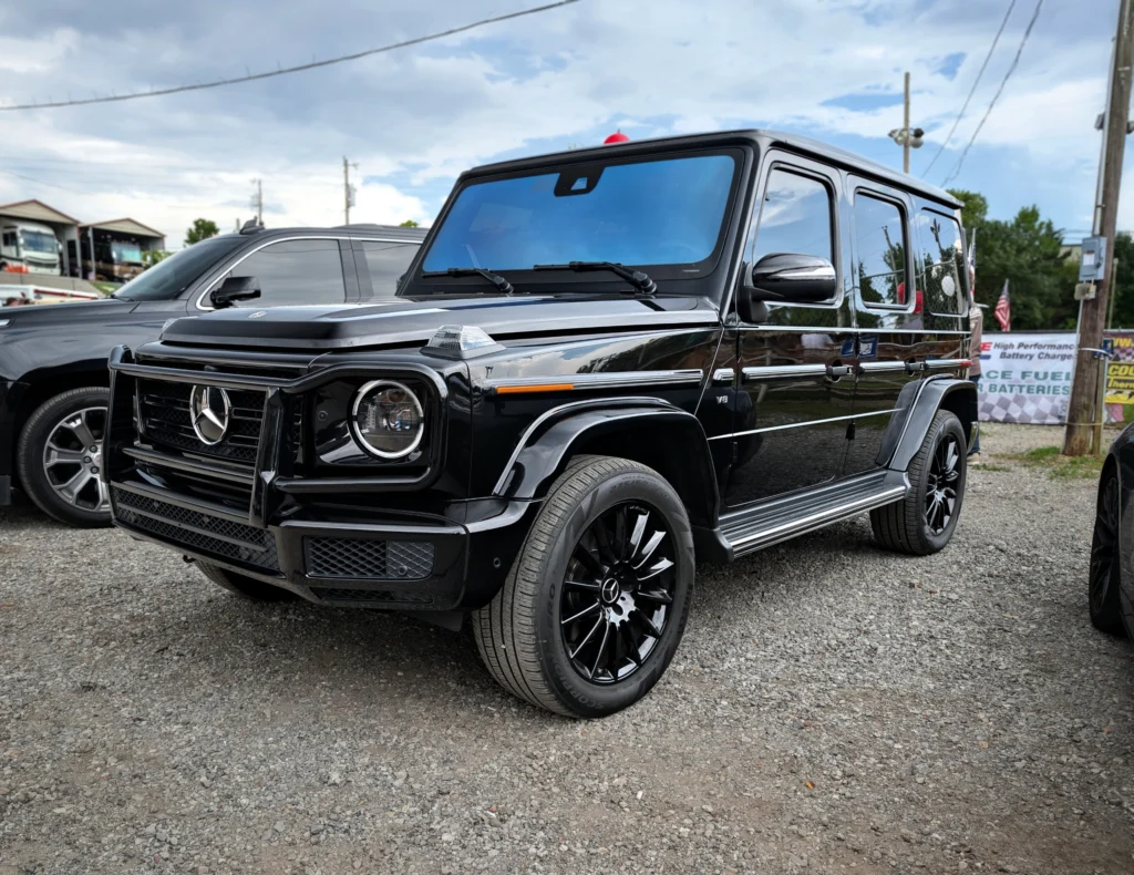 Black Mercedes-Benz G-Class SUV parked on gravel at an outdoor event with other vehicles and banners in the background