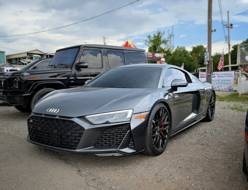 Gray Audi R8 and black Mercedes-Benz G-Class parked outdoors with utility poles and vendor tents in the background