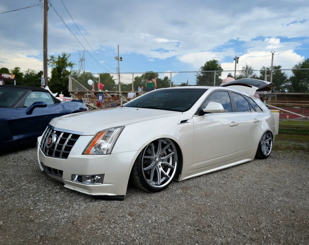 White Cadillac CTS Wagon with lowered suspension and aftermarket wheels parked on gravel at a car event with people and a chain-l