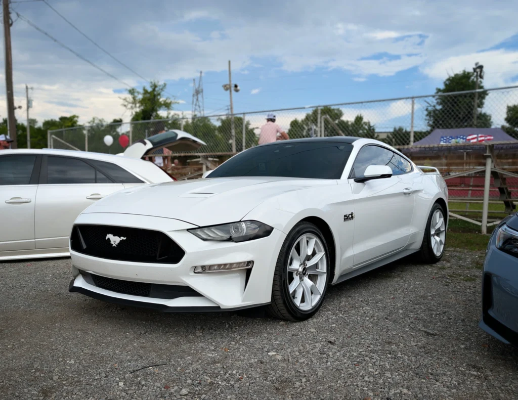 White Ford Mustang 5.0 parked on gravel at an outdoor car event with other vehicles and people in the background