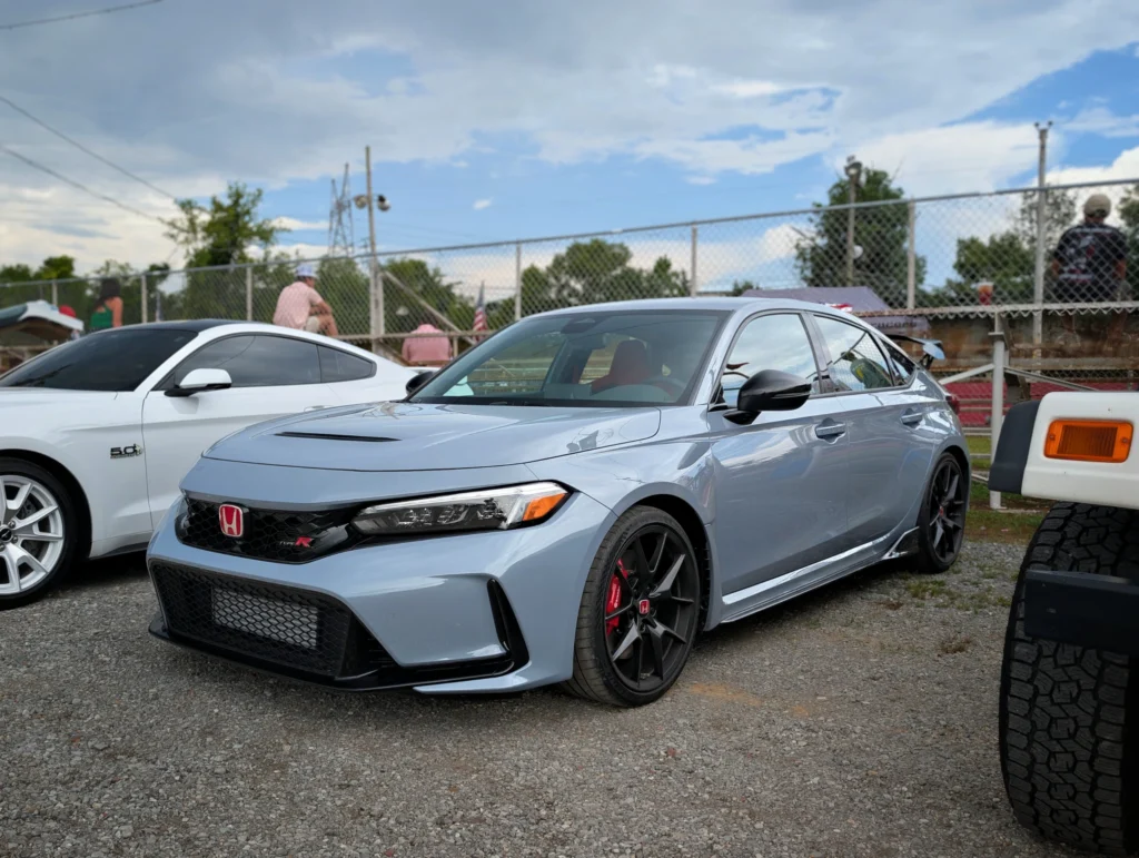 Gray Honda Civic Type R and white Ford Mustang GT parked on gravel at an outdoor car event with people seated behind a chain-