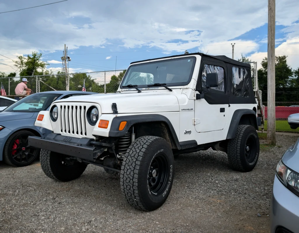White Jeep Wrangler with black soft top parked on gravel lot at car event with other cars and chain-link fence in background