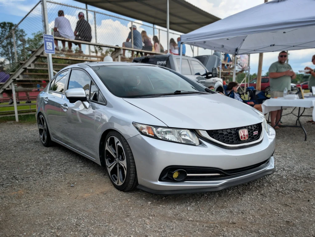 Silver Honda Civic Si sedan parked on gravel with people sitting on bleachers and under a canopy tent
