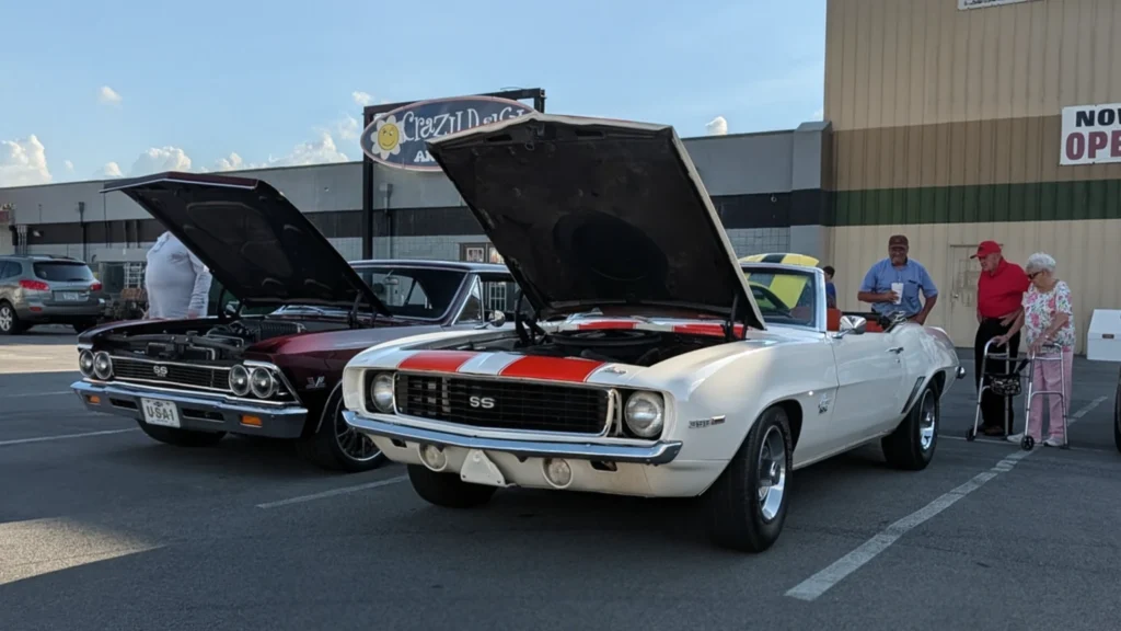 White Chevrolet Camaro SS convertible and maroon Chevrolet Chevelle SS with hoods open in a parking lot with people nearby