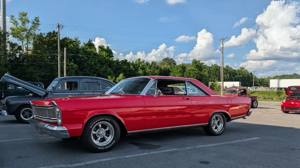 Red Ford Galaxie parked in a lot with other vintage cars under a blue sky with clouds