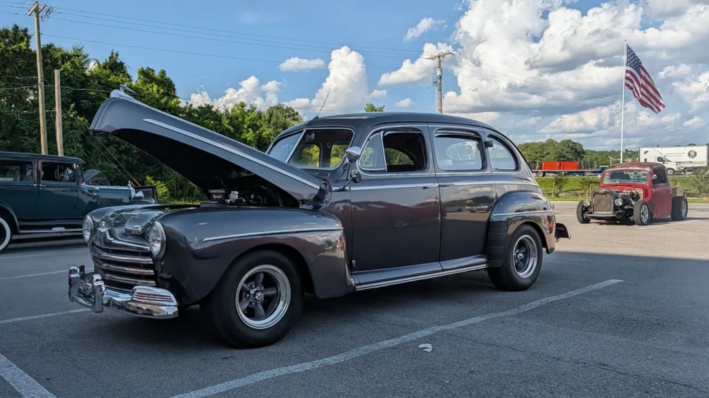 Dark gray 1946 Ford Super Deluxe four-door sedan with open hood parked outdoors at a car event with other vintage cars in bac