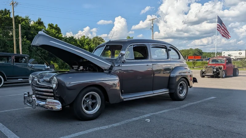 Dark gray 1946 Ford Super Deluxe four-door sedan with open hood parked outdoors at a car event with other vintage cars in bac