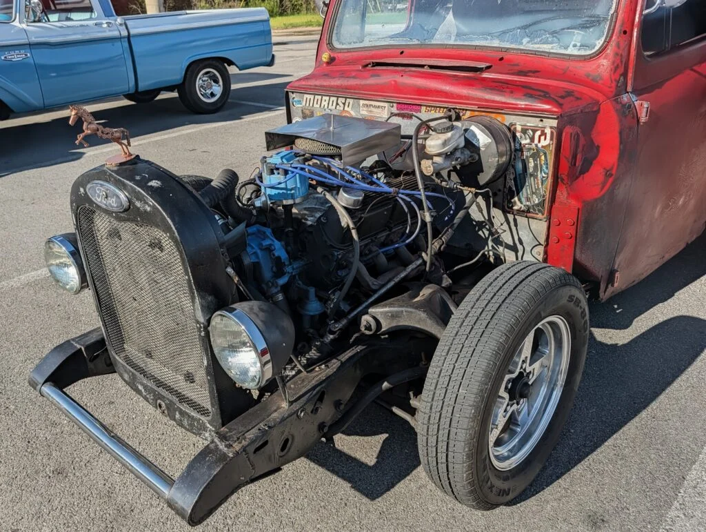Red rat rod Ford truck with exposed engine and blue distributor cap in a parking lot with a blue Ford pickup nearby