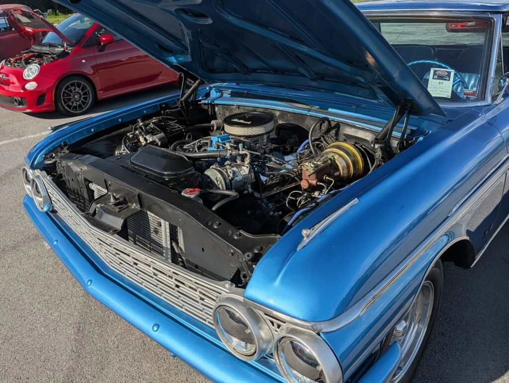 Blue vintage car with hood open showing engine, with red Fiat 500 Abarth in the background in a parking lot