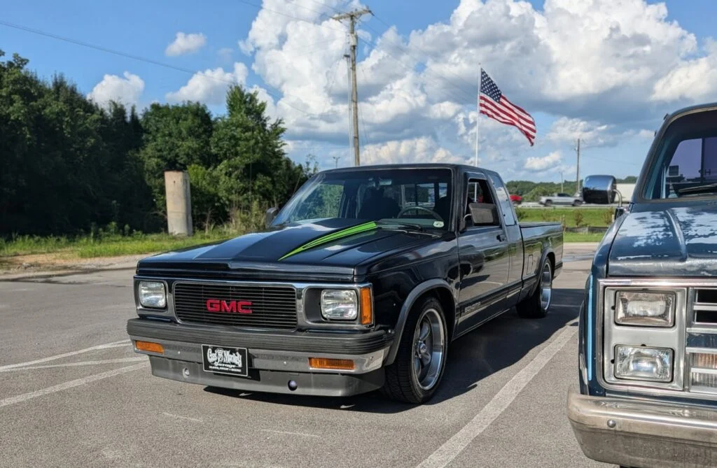 Black GMC Sonoma pickup truck parked next to a blue pickup in an outdoor lot with trees and an American flag in the backgroun