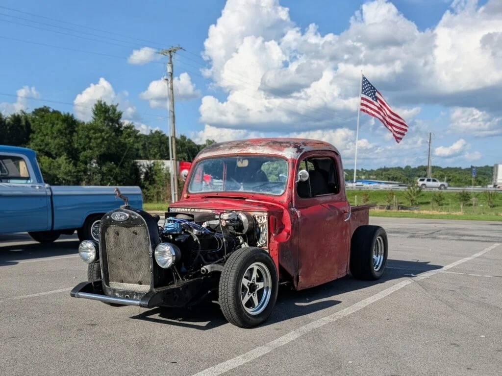 Red vintage Ford pickup truck in a parking lot with an American flag and a blue pickup in the background