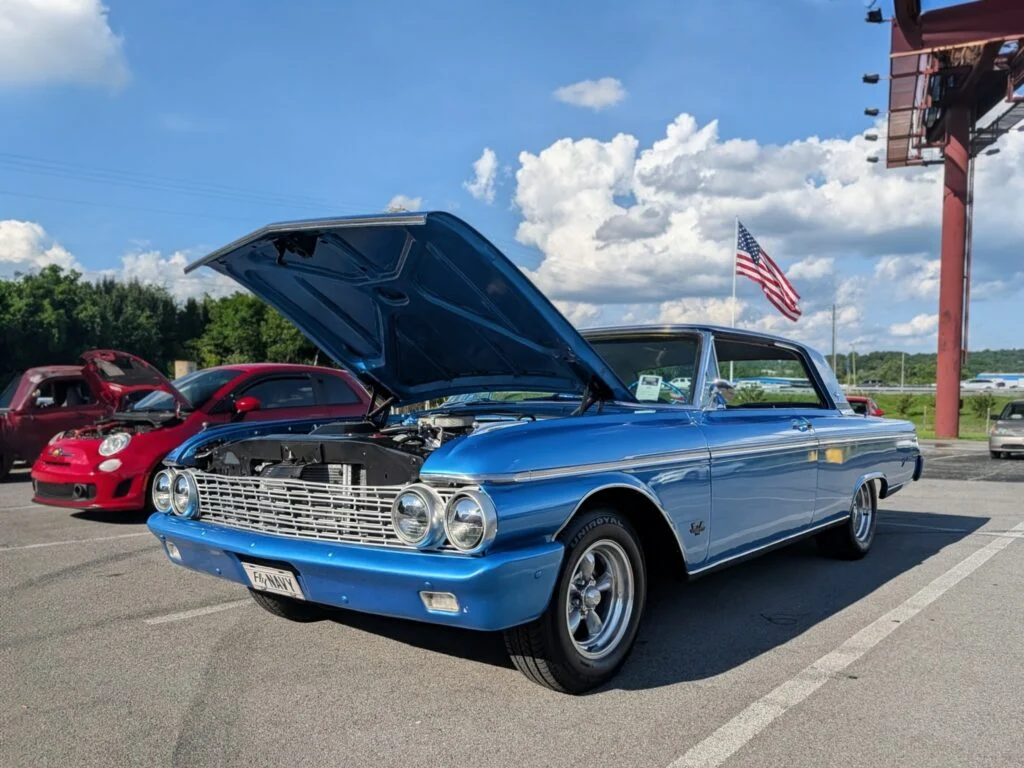 Blue Chevrolet Impala with hood open at an outdoor car event with American flag and other cars in background