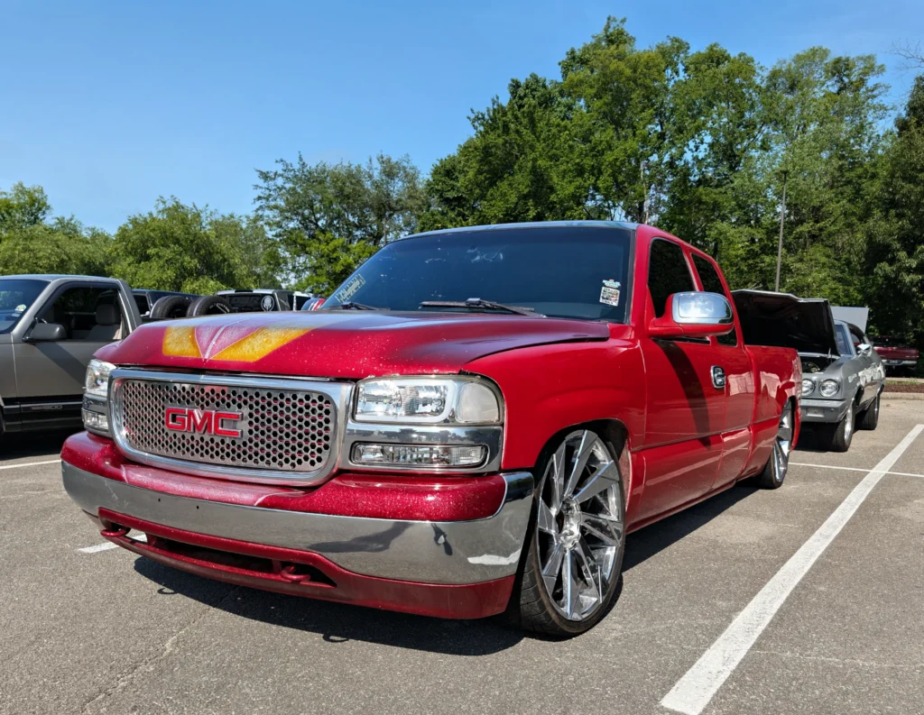 Red GMC pickup truck with custom paint and large chrome rims parked next to other vehicles in a parking lot with trees in the