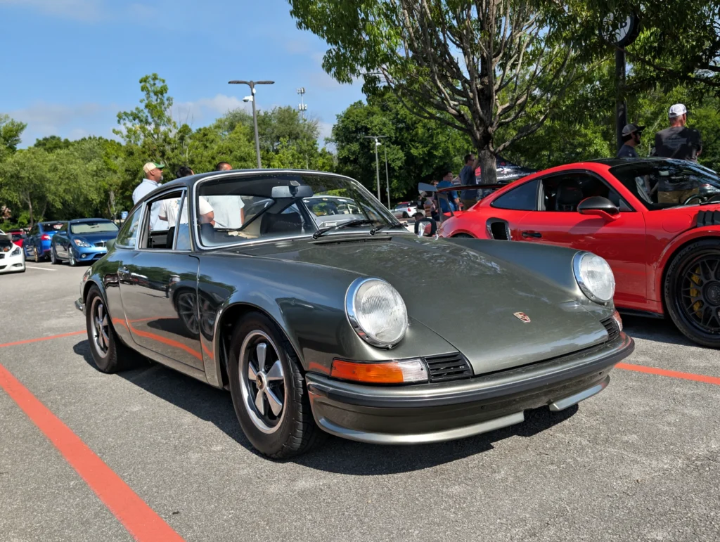 Gray Porsche 911 parked outside with several people and other cars nearby under a tree on a clear day.