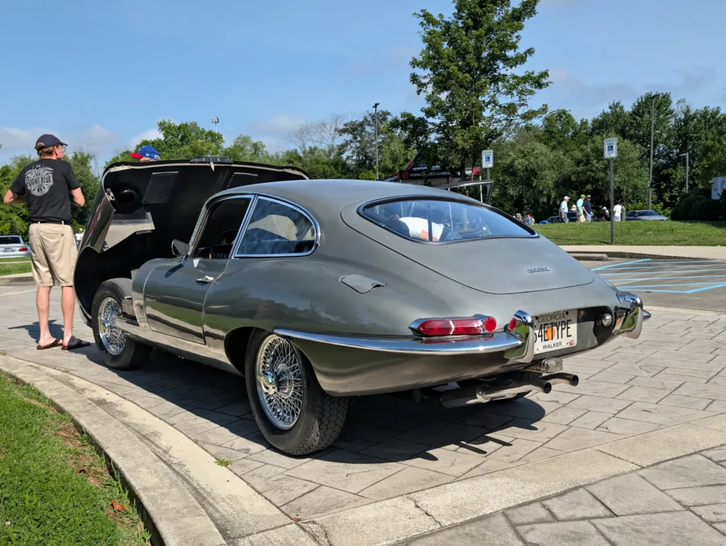 Silver Jaguar E-Type parked in an outdoor car event with its front hood open and two men standing nearby