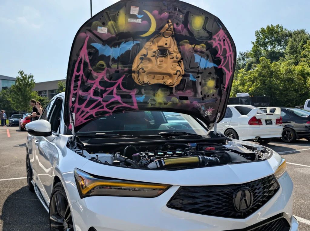 White Acura Integra with hood open showing painted underside at an outdoor car event parking lot