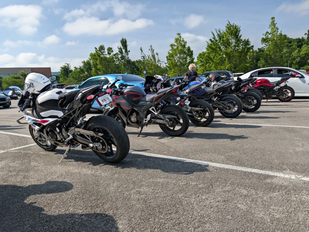 Five motorcycles parked in a row at an outdoor event parking lot with trees and people in the background