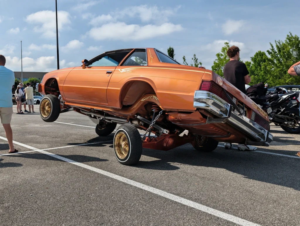 Orange Buick Regal with raised front wheels and gold rims at a car event parking lot with people and trees in the background
