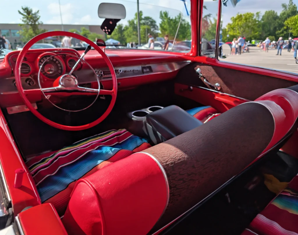 Interior view of a red car showing the dashboard, steering wheel, seats, and dashboard area with people and vehicles visible