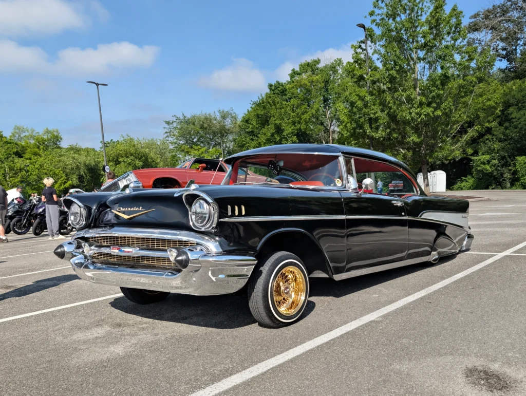 Black Chevrolet Bel Air with gold wheels parked at a car event on a sunny day with people and motorcycles in background