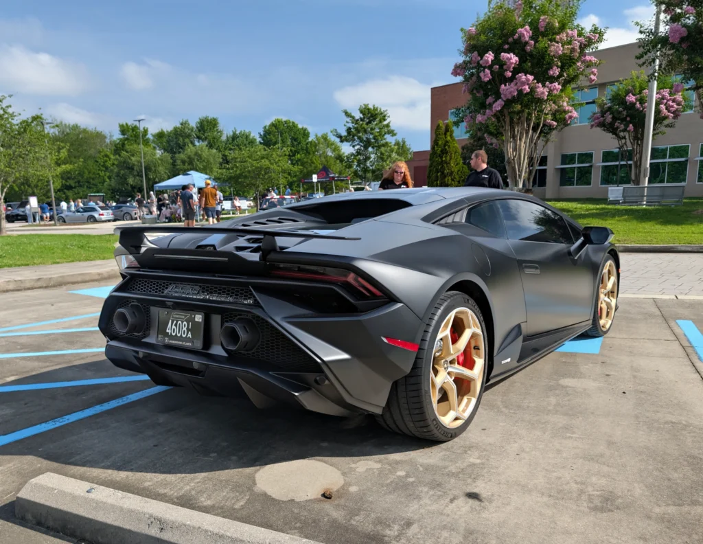 Matte black Lamborghini Huracán with gold wheels parked near people and tents at an outdoor car event under clear sky.