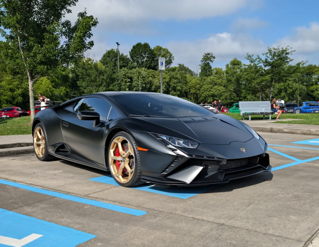 Matte black Lamborghini Huracan with gold wheels parked in a lot with trees and people in the background