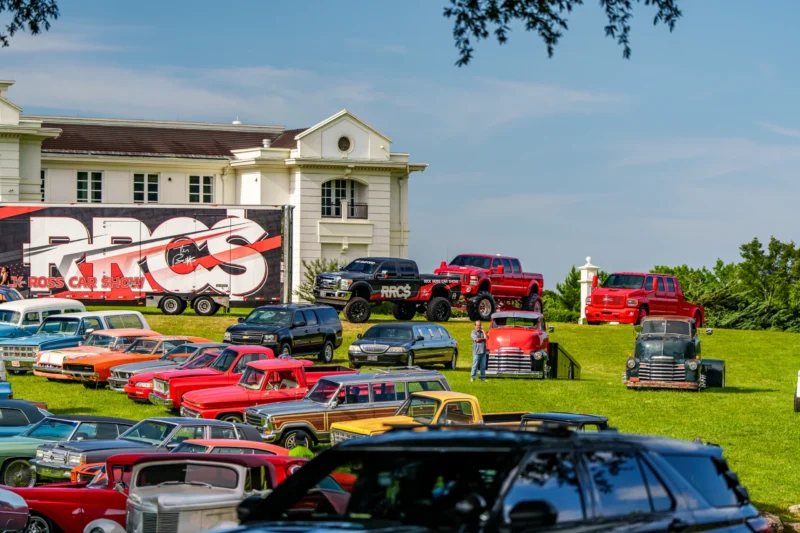 Multiple vintage and modern vehicles parked on a grassy hill at a car show with a large Rick Ross Car Show trailer and white