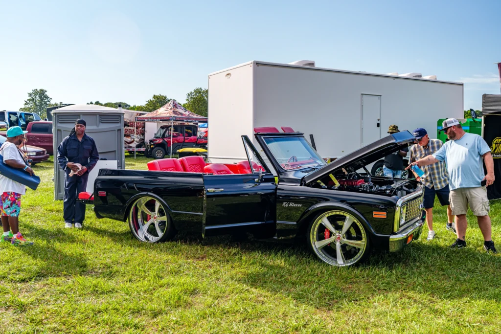 Black Chevrolet K5 Blazer convertible with red interior parked on grass at car event with several people nearby