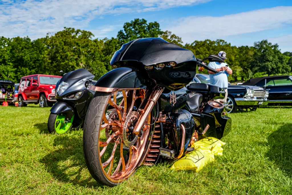 Two black motorcycles with copper components parked on grass, red SUV and classic car in the background under a blue sky.