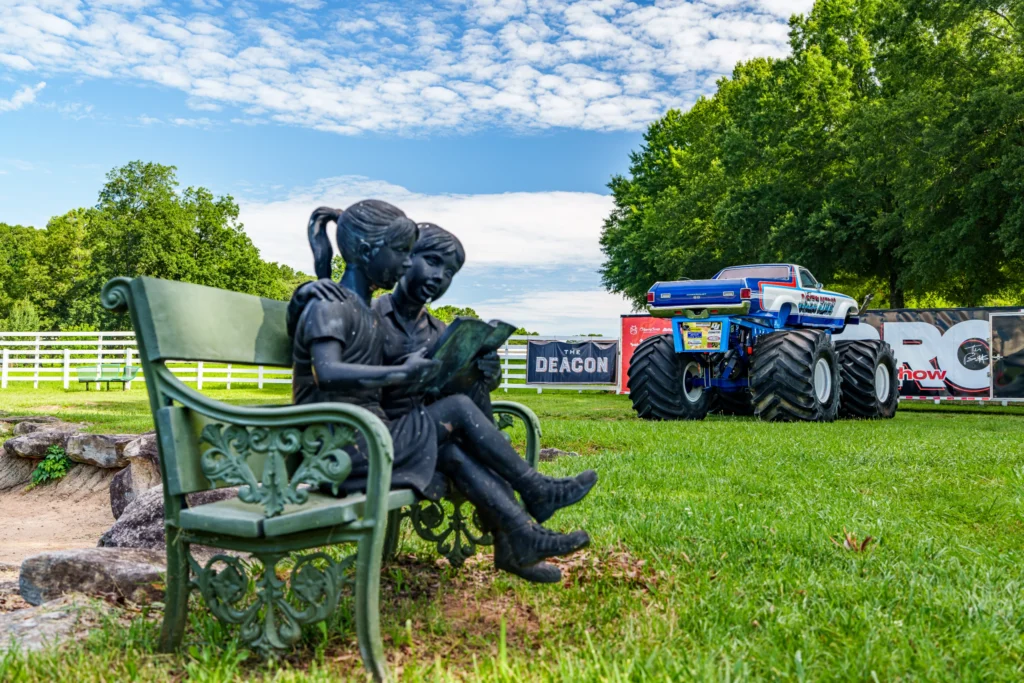 Blue Chevrolet El Camino SS monster truck parked on grass with trees and a statue of two children reading on a bench in foreg