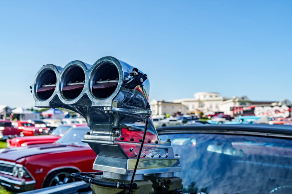 Close-up of a polished metal engine component with three circular openings at a car event with parked cars in the background