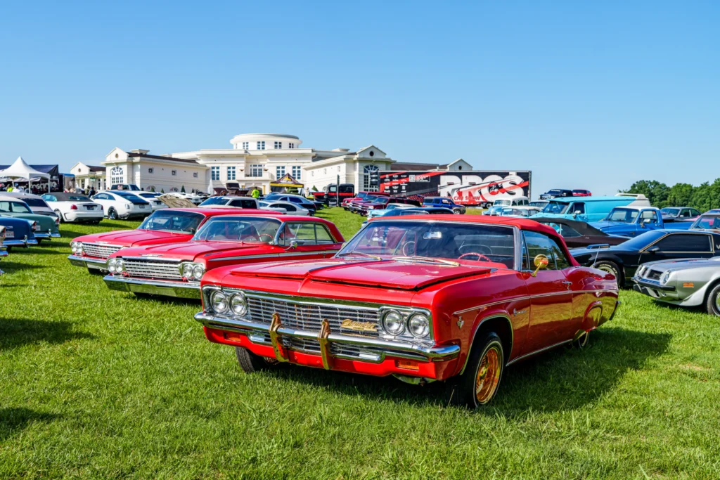 Red Chevrolet Impala in foreground at an outdoor car show on grass with a large white building in background under clear blue