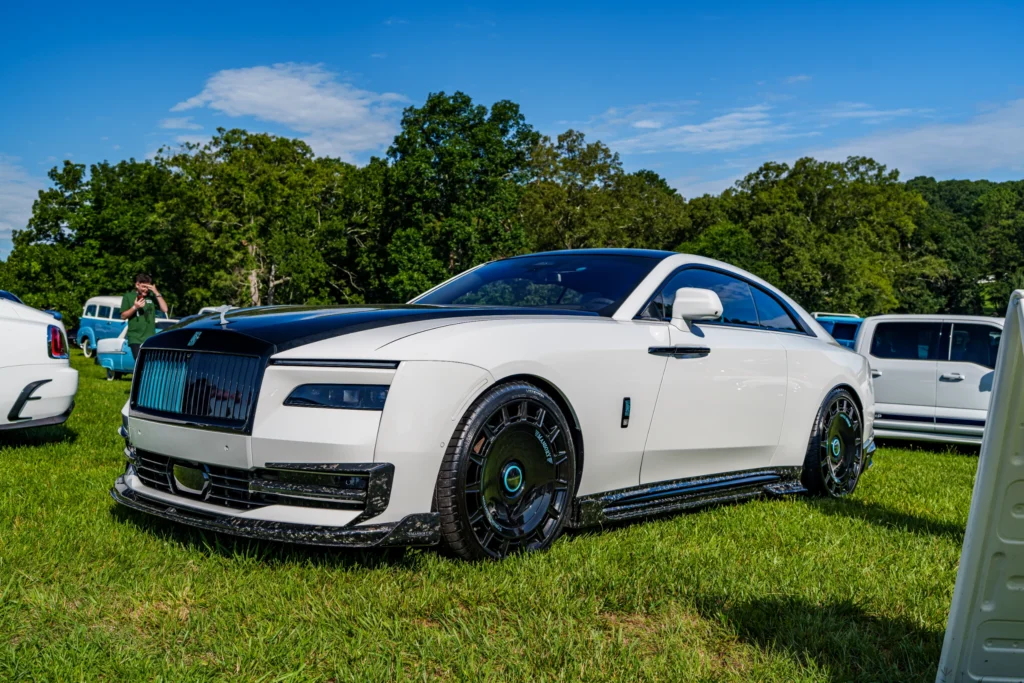 White Rolls-Royce coupe with black hood and custom wheels parked on grass with other vehicles and trees in background